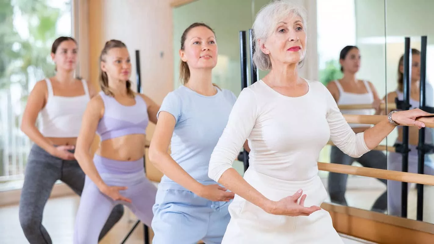 Slim aged woman and other attendees practicing ballet at ballet barre in dance studio during training session